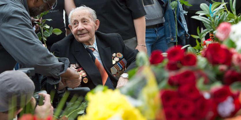 Ein alter Mann sitzt schräg hinter Rosen. Jemand schüttelt ihm die Hand