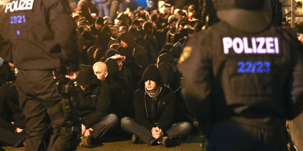 Zahlreiche Männer sitzen gefesselt vor Polizisten auf der Straße