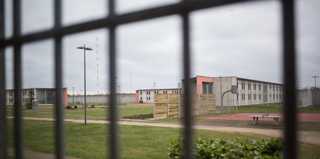 Durch die Gitterstäbe einer Gefängniszelle ist ein Glände mit niedrigen Häusern, Tischtennisplatte und Sportplatz zu sehen.