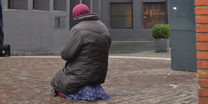 Eine Frau in einem dunklen Anorak und einem blauen Rock kniet vor einer Filiale der Bremischen Volksbank.