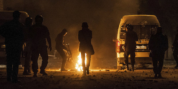 Mehrere uniformierte Polizisten um ein Feuer neben einem Auto in der Nacht