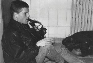 Zwei Männer auf dem Boden zwischen gekachelter Wand und Heizung, einer trinkt.