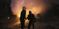 Zwei Männer, die Schutzhelme tragen, richten in der Dunkelheit einen Schlauch auf ein Feuer