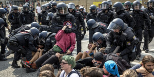 Schwarz gekleidete Polizeibeamte mit Helmen und Sturmhauben tragen Demonstranten von einer Straße. Ein Polizeibeamter hat einen Demonstranten im Würgegriff.