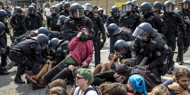 Schwarz gekleidete Polizeibeamte mit Helmen und Sturmhauben tragen Demonstranten von einer Straße. Ein Polizeibeamter hat einen Demonstranten im Würgegriff.