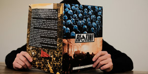 Ein Sammelalbum, auf dessen Cover Polizisten und ein Brand der G20-Proteste abgebildet sind. Das Heft einer schwarz gekleideten Person gehalten.