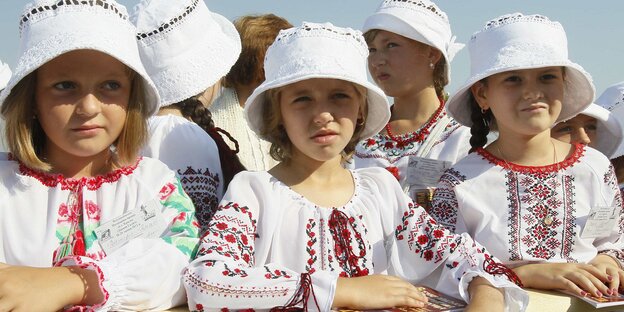 Mädchen mit weißen Huten und folkloristisch bestickten Blusen sitzen nebeneinander