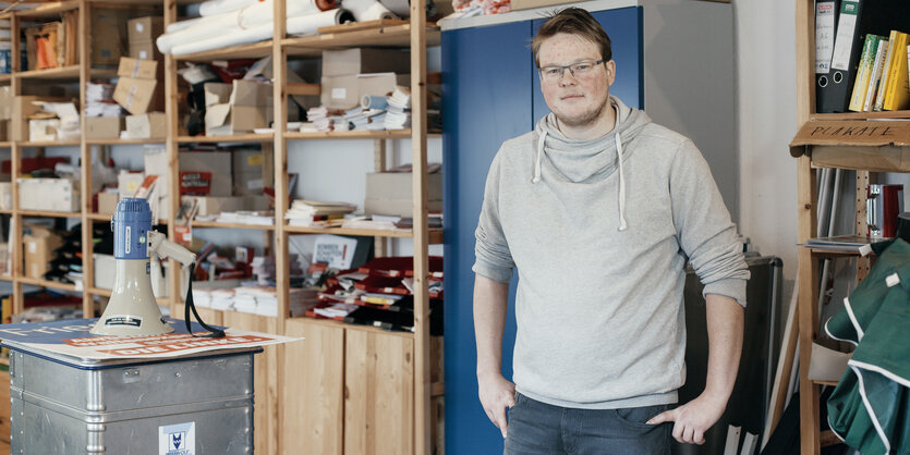 Ein junger Mann in grauem Pullover, im Hintergrund Holzregale, eine blaue Tür, neben ihm ein Schrank, auf dem ein Megafon ist