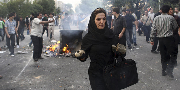 Eine Frau trägt Steine in der Hand. Hinter ihr brennen Blockaden