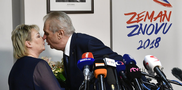 Ein Mann küsst eine Frau, vor ihm sind viele Mikrofone