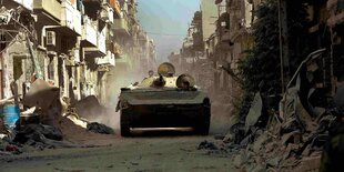 Ein Panzer fährt durch eine Straße in einer syrischen Stadt. An der Straßenseite stehen zerstörte Wohnhäuser.