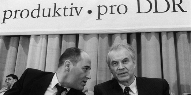 Der DDR Anwalt Gregor Gysi im Gespräch mit dem Vorsitzenden des DDR-Ministerrates, Hans Modrow. Im Hintergrund ist der Schriftzug "produktiv, pro DDR" zu sehen.