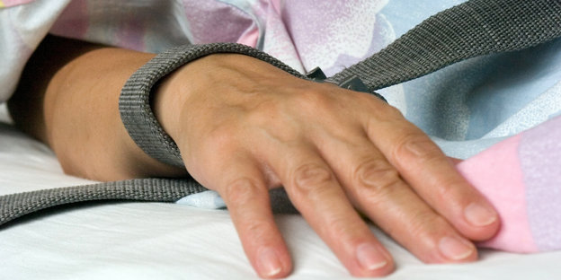 Eine Hand mit einem Band um das Handgelenk liegt auf einem Bett.