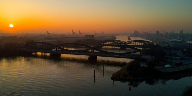 Die untergehende Sonne spiegelt sich in der Elbe. Es sind die Shiloutten der Elbbrücken und Container-Kräne zu sehen.