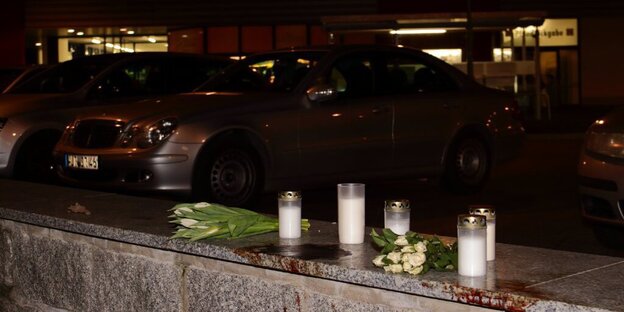 Kerzen und Blumen auf einem Mäuerchen, dahinter parkende Autos, dahinter ein Supermarkt