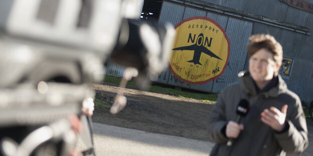Ein Reporter am Flughafen von Notre-Dame-des-Landes, der Schriftzug „Nein zum flughafen“ im Hintergrund