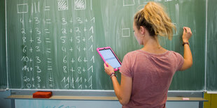 Eine Frau schreibt mit Kreide an eine Tafel und hält ein Tablet