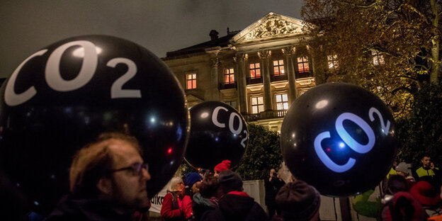 Menschen mit schwarzen Luftballons, auf denen „CO2“ steht vor einem erleuchteten Gebäude
