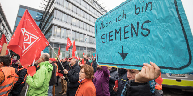 Demonstrationsteilnehmer mit Gewerkschaftsfahnen vor einem Haus, einer hält ein Schild hoch auf dem "Auch ich bin Siemens" steht