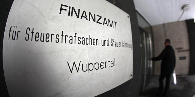 ein Schild mit der Aufschrift "Finanzamt für Steuerstrafsachen und Steuerfahndung - Wuppertal", im Hintergrund betritt ein Mann ein Gebäude