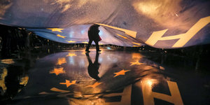 Ein Mensch verschwindet unter einer großen, blauen EU-Fahne, die sich über den Asphalt wölbt