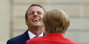 Macron lacht mit zurückgelegtem Kopf, Merkel steht davor