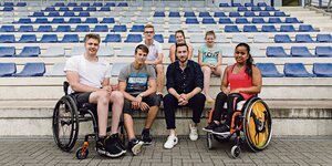Die sechs Nachwuchssportler*innen sitzen auf der Tribüne eines Stadions. Zwei von ihnn sitzen im Rollstuhl.