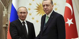zwei Männer schütteln sich die Hand, im Hintergrund eine russische und eine türkische Fahne