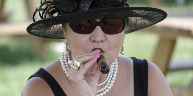 Ein Frau mit einem großen Hut und Perlenketten raucht eine Zigarre
