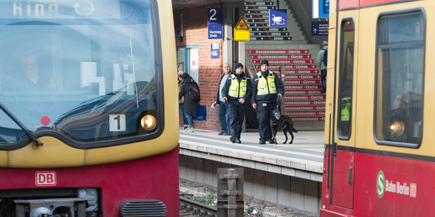 Zwei Sicherheitskräfte der Deutschen Bahn gehen in gelben Warnwesten mit einem Schäferhund über das Gleis, im Vordergrund fährt die Ringbahn.