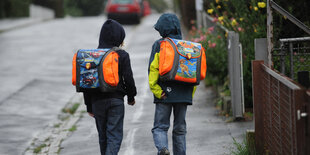 Zwei Schüler_innen, die Schulranzen tragen von hinten im Regen