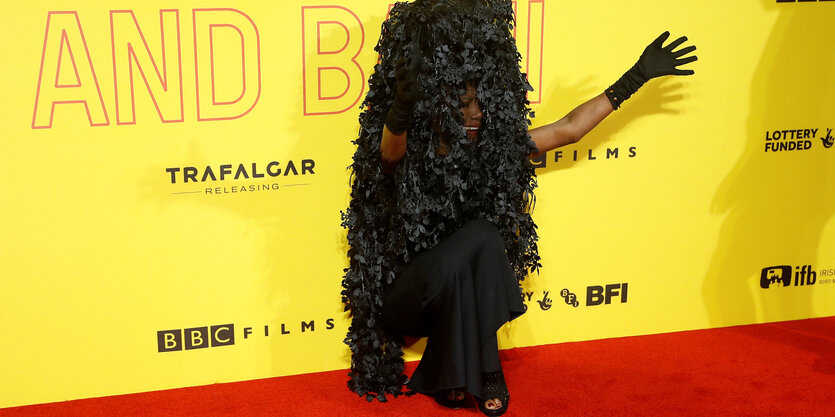 Grace Jones sitzt in einem schwarzen Kleid vor einer gelben Wand und streckt einen Arm aus