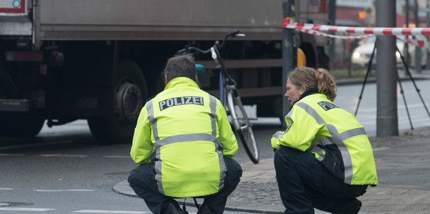 Zwei Polizisten in gelben Westen knien an einer Straßenkreuzung auf dem Boden. Im Hintergund steht ein Lastwagen.