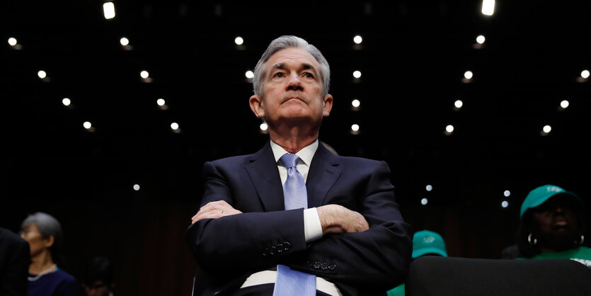 Jerome Powell sitzt mit verschränkten Armen, Lichterreihen an der Decke über ihm