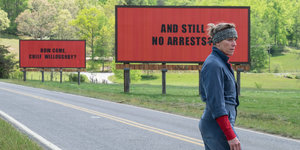 Eine Frau im Overall steht an einer Straße, im Hintergrund rote Werbetafeln mit schwarzer Aufschrift