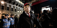 Jemand läuft mit einer Trumpmaske durch eine Stadt, im hintergrund stehen Polizisten