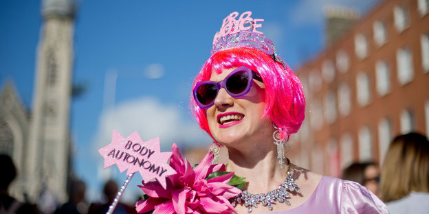 Eine Frau trägt eine pinkfarbene Perücke, Sonnenbrille und ein Krönchen. Sie hält ein Schild „Body Autonomy“ in die Höhe