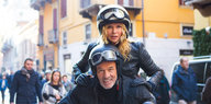 Ein Mann und eine Frau - beide mit Motorradhelm - schauen in die Kamera. Sie stützt sich auf ihn