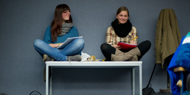 Zwei junge Frauen sitzen im Schneidersitz auf einem Tisch und halten Blöcke zum Mitschreiben.