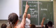 Kinder in einer Schulklasse melden sich. Der Lehrer steht vor einer beschriebenen Tafel.