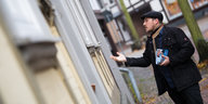 Ein junger Mann mit Bart und Schiebermütze steht an einer Haustür und gestikuliert mit der Hand.