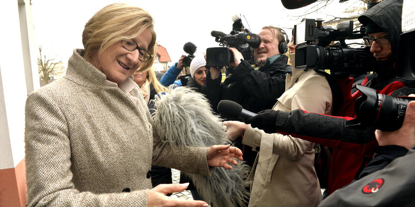 Eine Frau mit kurzen blonden Haaren gestikuliert, während sie vor vielen Mikrofonen spricht