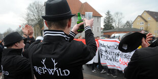 Drei Leute in schwarzen Kleidern mit dem Schriftzug "Freiwild" stehen vor einer Menschenmenge, die gegen das Konzert der Band demonstriert.