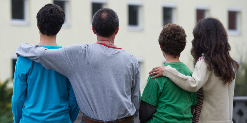 Ein Mann und eine Frau sind von hinten zu sehen, sie haben jeder den Arm um einen ihrer Söhne gelegt