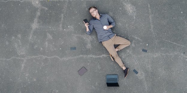 Jemand liegt auf dem Boden und schaut auf ein Smartphone um ihn herum liegen weitere Smartphones und ein Laptop