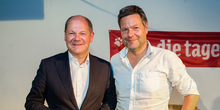 Ein älterer Mann mit Glatze und Sakko und ein jüngerer Mann im weißen Hemd stehen vor einem Banner der taz und lächeln.