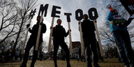 Vier schwarz gekleidete Menschen stehen auf einer Wiese und halten Schilder mit den Buchstaben von #MeToo hoch