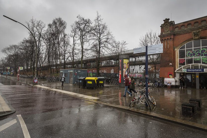 S-Bahn-Gleise auf einer Trasse führen in ein rotes Backsteingebäude. Davor ein breiter Gehsteig und eine Straße.