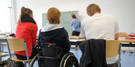 Drei Schüler sitzen in einem Klassenzimmer und schauen zur Tafel. Einer davon sitzt im Rollstuhl.