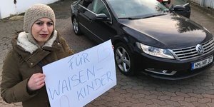 Eine junge Frau mit Kopftuch steht vor einem Auto und hält ein Schild mit der Aufschrift "Für Waisenkinder".